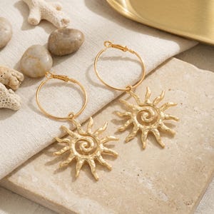 May include: Gold-colored hoop earrings with sunburst pendants. The sun pendants feature a spiral center and radiating rays. The earrings are displayed on a neutral-toned surface with decorative elements.