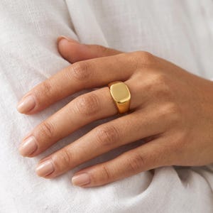 Könnte beinhalten: Ein polierter goldener Siegelring mit einer glatten, flachen, rechteckigen Oberfläche und einer massiven Ringschiene. Der Ring hat eine reflektierende, metallische Oberfläche und wird am Finger getragen, mit einem schlichten Design ohne Gravuren.