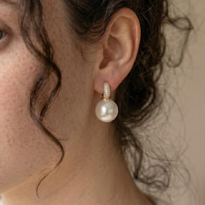 Op de afbeelding: Een enkele gouden oorring met een grote, witte parel hanger. De ring is bezet met kleine, heldere kristallen. De oorring wordt getoond op een oor, met de focus op de sieraden.
