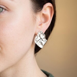 May include: A silver stud earring with an irregular, organic shape and a hammered, reflective surface texture. The earring features a polished metallic finish that catches the light, creating a series of small, uneven indentations across its face.