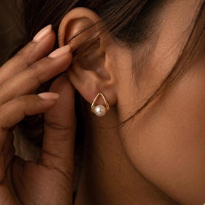 Op de afbeelding: Goudkleurige druppelvormige oorbellen met een witte parel in het midden. De oorbellen worden op het oor gedragen, waarbij de focus op de sieraden ligt. Het ontwerp is eenvoudig en elegant, geschikt voor dagelijks gebruik of speciale gelegenheden.