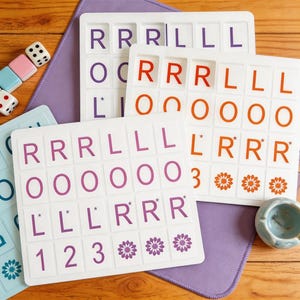 May include: White game boards with letter tiles in purple, orange, and red. The boards have letters and numbers, with flower designs. Dice and a small blue bowl are also visible.