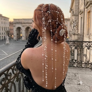 May include: A hair accessory featuring a delicate web of thin silver chains adorned with small white pearls that drape down the back. The piece is secured with a silver shell-shaped clip, accentuating a braided hairstyle with long, flowing strands.