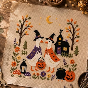 May include: A cross-stitch embroidery on beige fabric featuring two white geese wearing witch hats and capes. The scene includes a black house, a jack-o'-lantern, a cauldron, autumn trees, flowers, and a crescent moon. The text SnappyHue is visible.