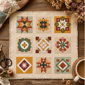 May include: A square piece of beige fabric featuring a cross-stitch pattern of nine distinct geometric blocks in shades of mustard yellow, sage green, dark red, and white. The text @Snappyhue is printed in white at the bottom center of the fabric.
