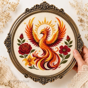 Könnte beinhalten: Eine kreisförmige Kreuzstichstickerei mit einem Phönix mit orangefarbenem, rotem und gelbem Gefieder unter einer Sonne. Das Design umfasst rote Rosen, eine gelbe Lilie und grüne Blätter auf cremefarbenem Stoff in einem verzierten bronzenen Rahmen.