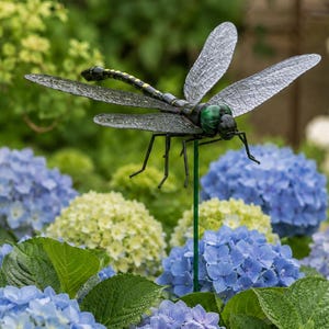 Pode incluir: Uma estaca de jardim de metal com uma libélula com asas translúcidas, cabeça verde e corpo preto. A libélula está posicionada acima de flores de hortênsia azuis e amarelas.