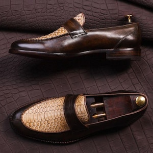 May include: Brown leather loafers with a snakeskin vamp and strap. The shoes have a classic design with a polished finish. The image shows two views of the shoes, one from the side and one from above, showcasing the details of the craftsmanship.