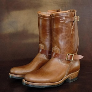 May include: A pair of mid-calf brown leather boots featuring a smooth finish, rounded toes, and stacked heels. Each boot is accented with a horizontal leather strap across the ankle and a vertical strap near the top, both secured with brass buckles.