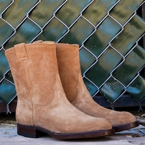 Könnte beinhalten: Ein Paar hellbraune Wildleder-Ankle Boots mit kleinem Absatz. Die Stiefel haben ein klassisches Design mit runder Spitze und einer Anziehschlaufe. Der Hintergrund zeigt einen grünen Maschendrahtzaun.