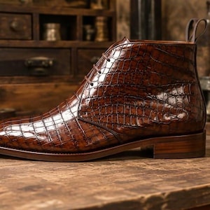 May include: A glossy dark brown ankle boot featuring a textured crocodile skin pattern. The boot has a lace-up front with thin brown laces, a stacked wooden heel, and a pull tab at the back collar. The finish is highly polished and reflective.