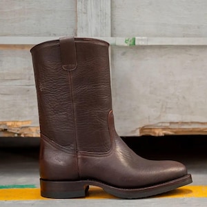 Peut inclure: Botas de cowboy en cuir marron, mi-mollet. La botte a un bout arrondi, un petit talon et une tirette. Le cuir a un aspect texturé et une couleur marron foncé. La botte mesure environ 30 cm de haut.