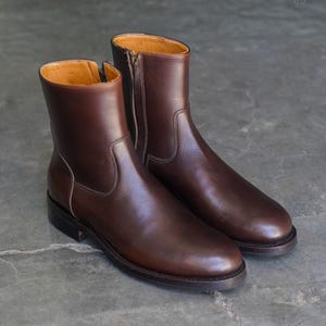 May include: A pair of brown leather ankle boots with side zips. The boots have a smooth, polished finish and a light brown interior. The soles are dark brown with a subtle heel. The boots are displayed on a grey surface.