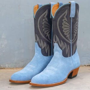 May include: A pair of light blue suede cowboy boots. The boots have a dark gray shaft with decorative stitching and a light blue pull tab. The soles and heels are brown. The interior is a light orange color.