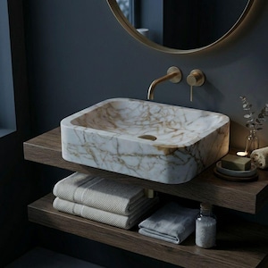 May include: A rectangular marble sink with gold veining sits on a wooden shelf. Below, folded towels and a small bottle of bath salts are visible. A gold faucet and round mirror are mounted on the dark gray wall, creating a spa-like bathroom setting.