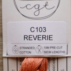 May include: A skein of coral-colored stranded cotton embroidery thread on a branded card labeled C103 REVERIE. The packaging specifies 10M pre-cut 50cm lengths of cotton thread. The card features a circular logo with a stylized fan design.