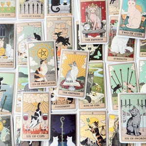 May include: A collection of tarot cards featuring various cat illustrations. Each card displays a unique feline character in different settings, including The Emperor, The Empress, Six of Cups, and Six of Swords, with titles printed at the bottom.