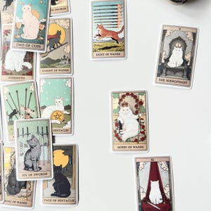 May include: A collection of rectangular tarot cards featuring various cat illustrations. Each card displays a unique feline character in different settings, including The Hierophant, Queen of Wands, Two of Cups, Eight of Wands, and Six of Swords.
