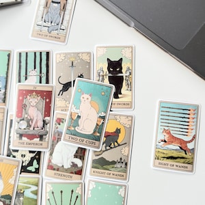 May include: A collection of rectangular tarot cards featuring various cat illustrations. Visible cards include The Emperor, Two of Cups, Strength, Knight of Wands, Eight of Wands, Two of Swords, and Ace of Swords. Each card displays a unique feline character.