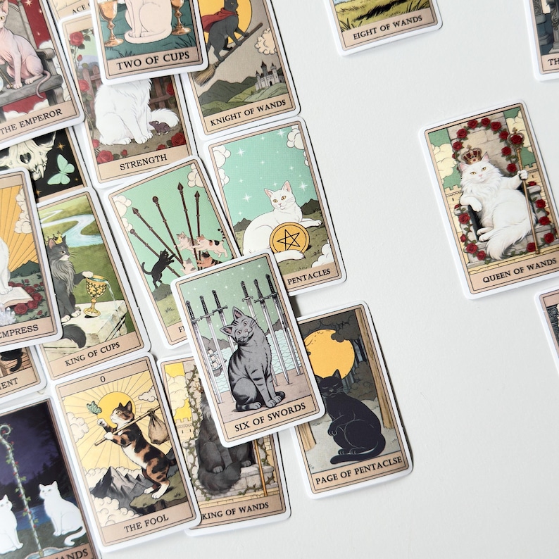 May include: A collection of rectangular tarot cards featuring illustrations of various cat breeds in different settings. Each card displays a unique scene with a cat, including titles like The Emperor, Strength, Queen of Wands, and Six of Swords.