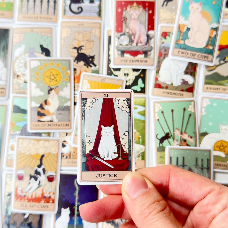 May include: A rectangular Justice tarot card sticker featuring a white cat with a blindfold sitting in front of a red curtain, holding a sword and scales. The card is bordered in cream with the Roman numeral XI and the text JUSTICE at the bottom.