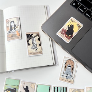 May include: A collection of rectangular stickers featuring cat-themed tarot card illustrations. Designs include The Magician, The Hanged Man, Six of Pentacles, and Two of Swords. The stickers are displayed on an open grid notebook and a laptop surface.