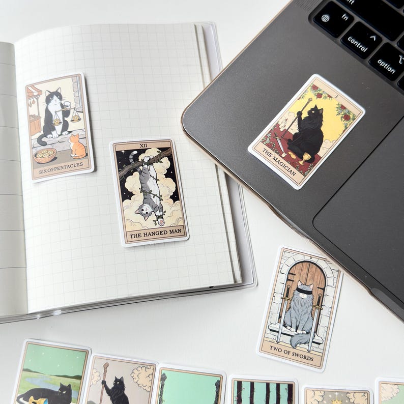 May include: A collection of rectangular stickers featuring cat-themed tarot card illustrations. Designs include The Magician, The Hanged Man, Six of Pentacles, and Two of Swords. The stickers are displayed on an open grid notebook and a laptop surface.