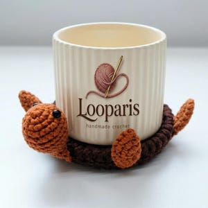 May include: A handmade crochet turtle coaster in shades of brown and orange, with a cream-colored mug. The mug has a logo that says "Looparis handmade crochet". The coaster is designed to hold the mug.