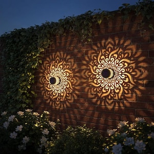 May include: Two solar-powered wall lights with intricate, mandala-like patterns casting decorative shadows on a brick wall. The lights are circular with a crescent-shaped solar panel. Green foliage and white flowers are visible.