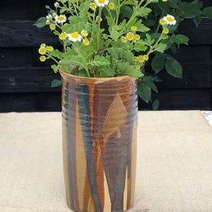 May include: A cylindrical ceramic vase with a glossy glaze featuring vertical streaks of brown, tan, and dark blue. The vase is filled with a bouquet of white and yellow daisy-like flowers with green stems and leaves, standing upright.