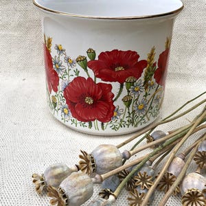 May include: A white ceramic planter featuring a vibrant botanical print of red poppies, small blue and white wildflowers, and green wheat stalks. The cylindrical vessel has a smooth finish and a thin gold-toned rim along the top edge.