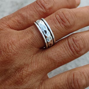 May include: A silver spinner ring featuring a wide, hammered center band with a textured, dimpled surface, flanked by two smooth, polished silver bands on either side. The metal has a bright, reflective finish throughout the entire ring design.