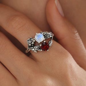 Könnte beinhalten: Ein Silberring mit einem runden, schillernden Mondstein und einem runden, tiefroten Granat-Edelstein. Die Ringschiene ist mit detaillierten Metallarbeiten verziert, die Blätter und eine kleine Blume darstellen und eine organische, texturierte Optik erzeugen.