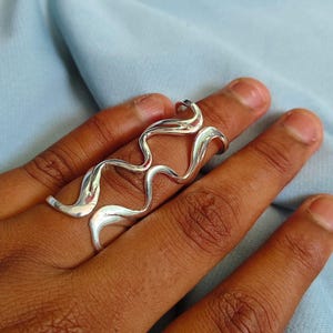 May include: A silver-colored ring with a flowing, organic design. The ring wraps around two fingers, featuring a series of curved, wave-like shapes. The jewelry is displayed against a light blue fabric background.