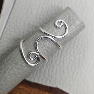 Puede incluir: Un anillo plateado con un diseño abierto y ondulado. El anillo se envuelve alrededor del dedo con una línea continua y fluida, creando un aspecto único y artístico. El anillo se muestra sobre una superficie gris.
