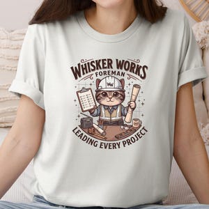 May include: A light-colored t-shirt featuring a cartoon cat in a construction worker outfit. The cat is holding a clipboard and blueprints, with the text "Whisker Works Foreman Leading Every Project" above and below the image.