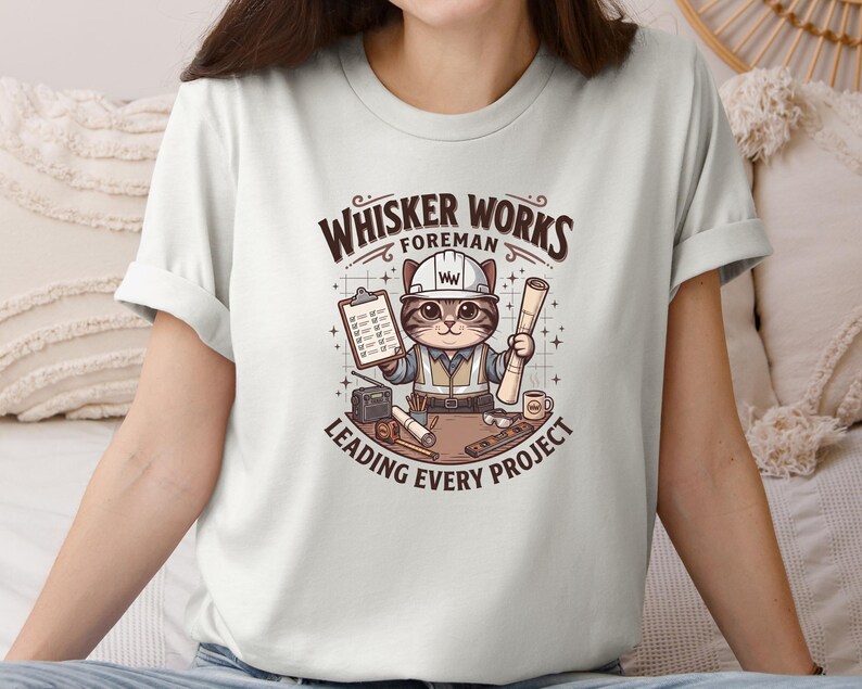 May include: A light-colored t-shirt featuring a cartoon cat in a construction worker outfit. The cat is holding a clipboard and blueprints, with the text "Whisker Works Foreman Leading Every Project" above and below the image.