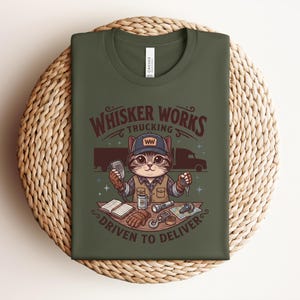 May include: Dark green t-shirt with a cartoon cat trucker design. The text reads "Whisker Works Trucking" and "Driven to Deliver". The cat is depicted with a cap, microphone, and keys, alongside a truck and other trucking-related items.