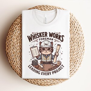 May include: White t-shirt featuring a cartoon cat in a construction worker outfit, holding blueprints and a clipboard. The text reads "Whisker Works Foreman Leading Every Project". The design includes a hard hat, tools, and a coffee mug.