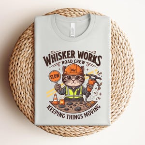 May include: Pale grey t-shirt featuring a cartoon cat in a construction worker's outfit. The cat holds a "SLOW" sign and a jackhammer. Text reads "WHISKER WORKS ROAD CREW" and "KEEPING THINGS MOVING".