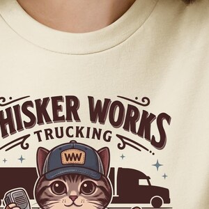 May include: A cream-coloured t-shirt with a cartoon cat wearing a trucker hat with "WW" on it. The text "WHISKER WORKS TRUCKING" is arched above the cat, with truck and trailer graphics. The cat is holding a walkie-talkie.
