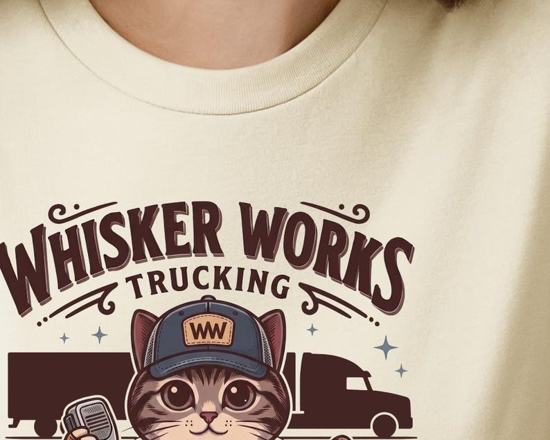 May include: A cream-coloured t-shirt with a cartoon cat wearing a trucker hat with "WW" on it. The text "WHISKER WORKS TRUCKING" is arched above the cat, with truck and trailer graphics. The cat is holding a walkie-talkie.