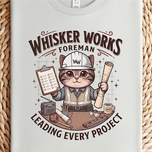 May include: A light gray t-shirt featuring a cartoon cat dressed as a foreman. The cat wears a hard hat and holds a clipboard and rolled blueprints. Text reads "Whisker Works Foreman Leading Every Project."