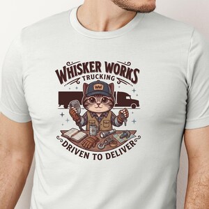 May include: White t-shirt with a cartoon cat trucker design. The design includes the text "Whisker Works Trucking" and "Driven to Deliver." The cat is wearing a cap and holding a microphone and keys. A lorry is in the background.