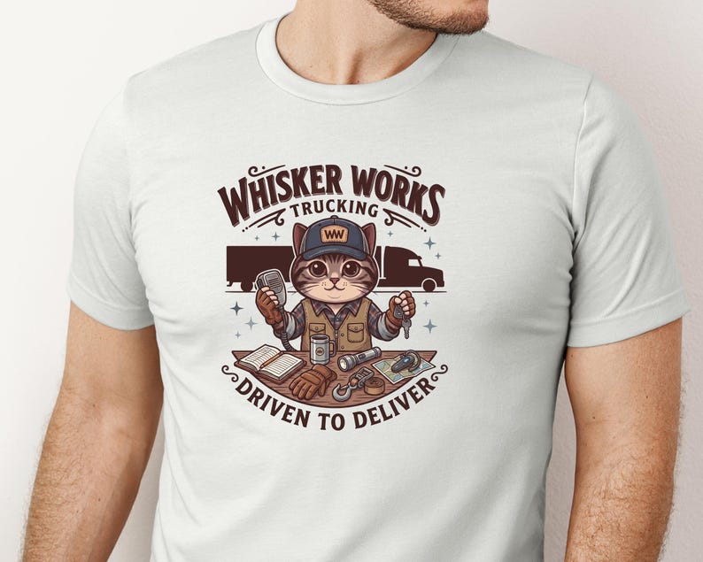 May include: White t-shirt with a cartoon cat trucker design. The design includes the text "Whisker Works Trucking" and "Driven to Deliver." The cat is wearing a cap and holding a microphone and keys. A lorry is in the background.