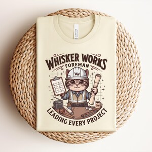 May include: Cream-colored t-shirt featuring a cartoon cat foreman wearing a hard hat and holding blueprints and a clipboard. The text reads "Whisker Works Foreman Leading Every Project."