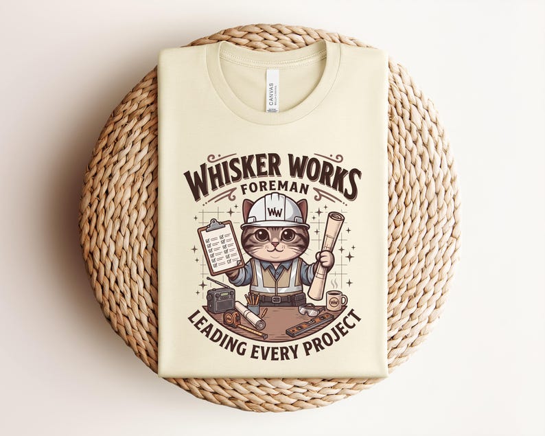 May include: Cream-colored t-shirt featuring a cartoon cat foreman wearing a hard hat and holding blueprints and a clipboard. The text reads "Whisker Works Foreman Leading Every Project."
