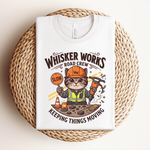 May include: White t-shirt with a cartoon cat in a construction worker outfit, holding a "SLOW" sign and a jackhammer. The shirt has the text "WHISKER WORKS ROAD CREW KEEPING THINGS MOVING". The design includes an orange traffic cone and a flashing light.