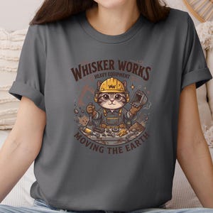 May include: A charcoal gray t-shirt featuring a cartoon cat wearing a yellow hard hat and overalls, holding tools. The shirt has the text "Whisker Works Heavy Equipment Moving the Earth" in a brown, distressed font. The cat is surrounded by construction tools.