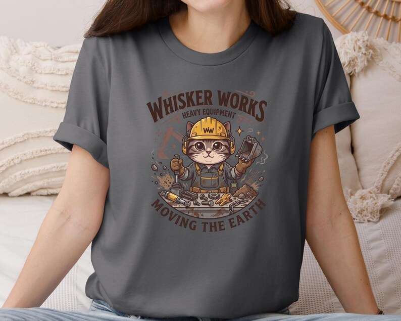 May include: A charcoal gray t-shirt featuring a cartoon cat wearing a yellow hard hat and overalls, holding tools. The shirt has the text "Whisker Works Heavy Equipment Moving the Earth" in a brown, distressed font. The cat is surrounded by construction tools.
